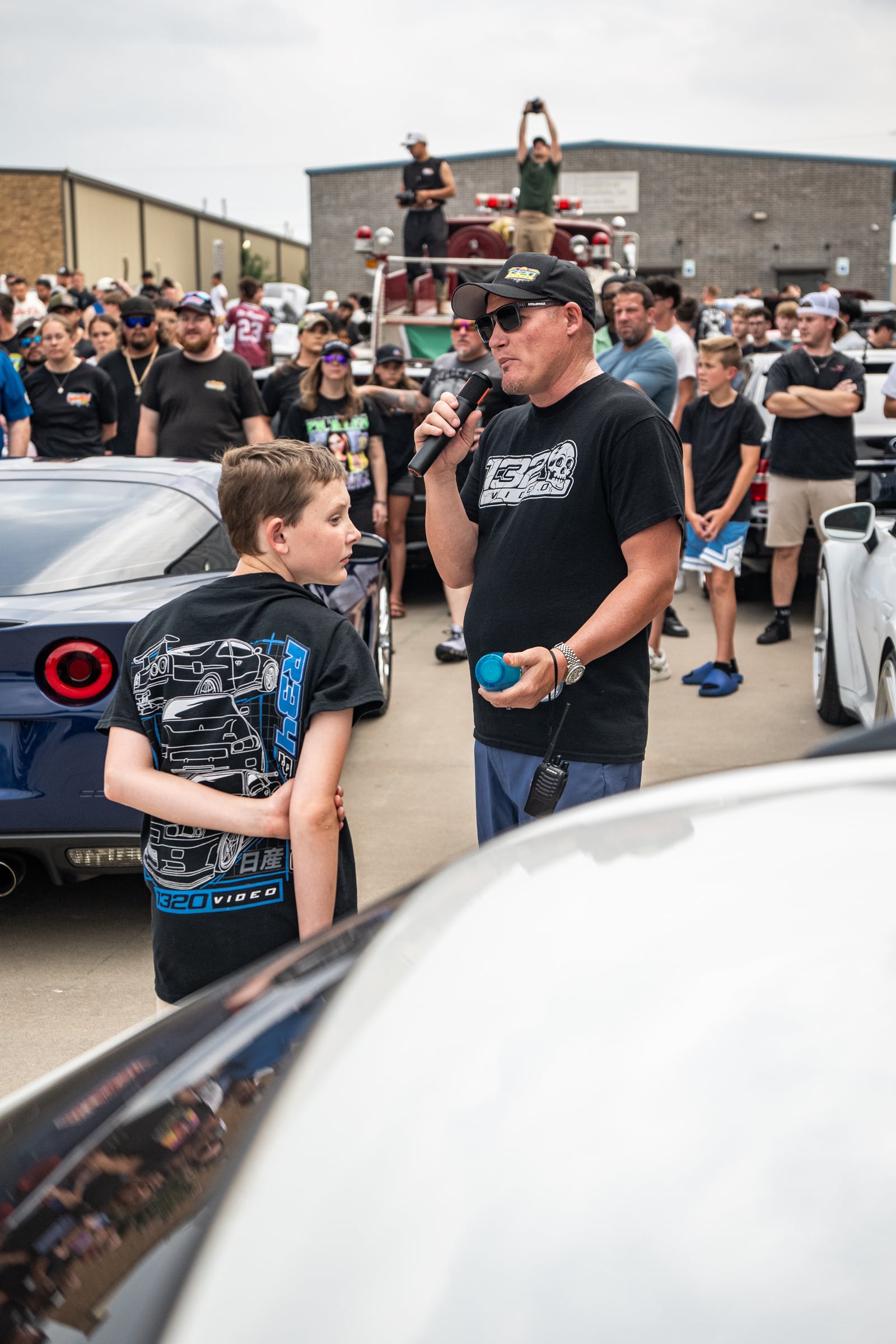 A speaker addresses the memorial cruise crowd while a young attendee wears a 1320Video shirt.